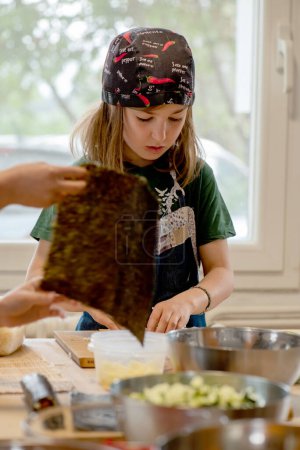Photo for Girl making sushi rolls, dressed in a Texan apron and chef's hat. Hair tousled outside, in front out of focus another girl's hands with a nori seaweed. - Royalty Free Image