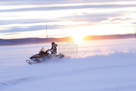 Active woman is driving a snowmobile on the snowy field in the sunset time.