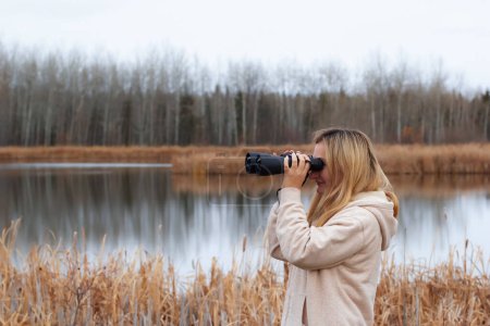 A blond caucasian woman in beige coat is watching through binoculars in the park near the lake in late autumn.
