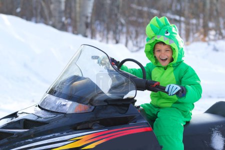 Photo for Ten year old boy is sitting on a black snowmobile in a cool bright green dragon-like snowsuit  and laughing in the winter forest. - Royalty Free Image