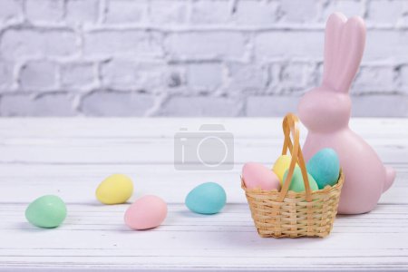 Foto de Composición mínima de Pascua, estandarte con figurita rosa de un conejo y una pequeña canasta de mimbre con coloridos caramelos en forma de huevo. - Imagen libre de derechos