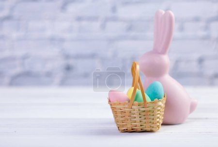 Foto de Composición mínima de Pascua, estandarte con figurita rosa de un conejo y una pequeña canasta de mimbre con coloridos caramelos en forma de huevo. - Imagen libre de derechos