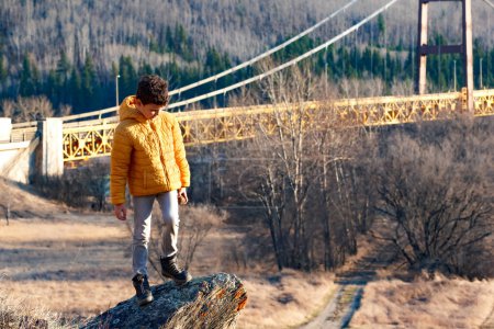 Scenic spring landscape with bare trees and dry yellow grass, a boy in yellow jacket is standing on a rock on the hill with view to the bridge. Dunvegan bridge, Alberta, Canada.