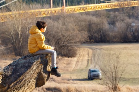 Scenic spring landscape with bare trees and dry yellow grass, a boy in yellow jacket is sitting on a rock on the hill and looking at the bridge. Dunvegan bridge, Alberta, Canada.
