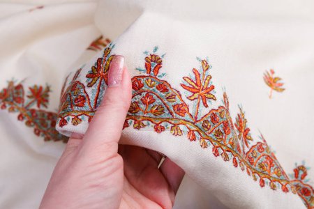 Schöner indischer Schal, Kaschmirstickerei auf Wolle. Hand hält weißen Schal mit goldenem und braunem traditionellen Muster.