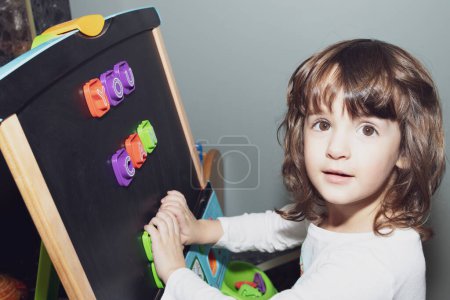 Cute little girl is learning letters at home on her art easel. Colorful magnetic letters say "You can".