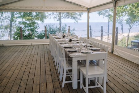 Téléchargez les photos : Une table couverte de blanc pour un événement sous un auvent blanc sur le bord de la mer - en image libre de droit