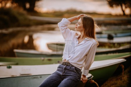 Téléchargez les photos : Une femme est assise au coucher du soleil en jeans et une chemise blanche, sur le bord d'un lac - en image libre de droit