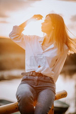 Téléchargez les photos : Une femme est assise au coucher du soleil en jeans et une chemise blanche, sur le bord d'un lac - en image libre de droit