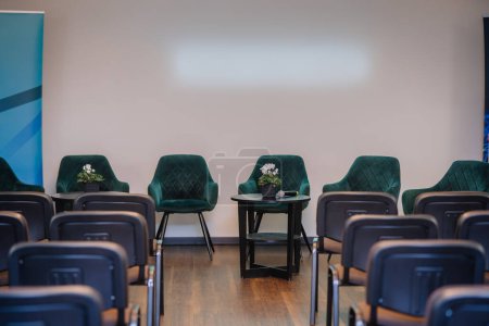  installation simple d'un séminaire avec une ligne de fauteuils luxueux en velours vert derrière une petite table ronde, une plante en pot sur le dessus et des bannières roll-up graphiques bleues sur le côté.