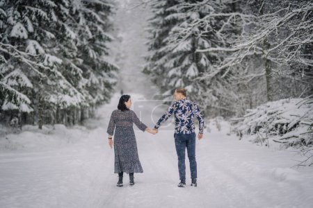 man and woman holding hands while walking on a snow-covered path through a forest, looking back towards the camera.