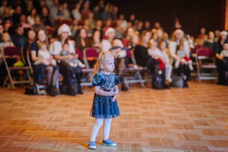 Téléchargez les photos : Valmiera, Lettonie - 28 décembre 2023 - jeune fille en robe bleue étincelante avec des motifs étoilés se tient seul au premier plan lors d'une performance, face à un public qui comprend des enfants portant des chapeaux de Père Noël - en image libre de droit
