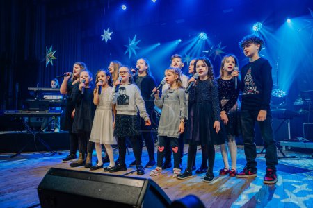 Téléchargez les photos : Valmiera, Lettonie - 28 décembre 2023 - groupe d'enfants dans diverses tenues chante dans des microphones sur une scène décorée avec des motifs étoilés et éclairage de scène bleu. - en image libre de droit