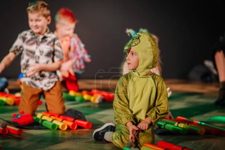 Photo for Valmiera, Latvia - December 28, 2023 - child in a green dragon costume sitting on the floor among colorful toy instruments, watching other children play in a blurred background. - Royalty Free Image