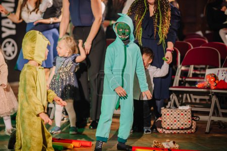 Foto de Valmiera, Letonia - 28 de diciembre de 2023 - los niños están vestidos con trajes coloridos para una actuación o un juego, destacando uno con un atuendo verde. Están en una sala con una audiencia en el fondo. - Imagen libre de derechos