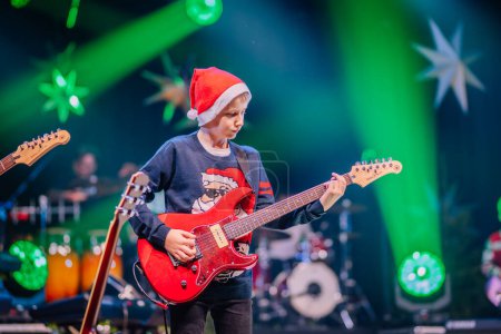 Photo for Valmiera, Latvia - December 28, 2023 - young boy in a Santa hat playing a red electric guitar on stage with green stage lighting and star decorations in the background - Royalty Free Image