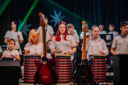 Foto de Valmiera, Letonia - 28 de diciembre de 2023 - grupo de niños con camisas blancas enfocados en tocar instrumentos de percusión en el escenario, con guitarras visibles y un telón de fondo estrellado. - Imagen libre de derechos