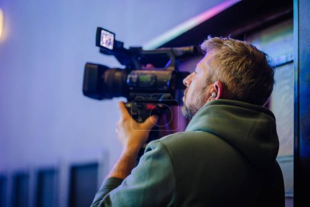 Foto de Camarógrafo que opera una cámara de vídeo profesional, se centró en su tarea con un fondo colorido escenario - Imagen libre de derechos