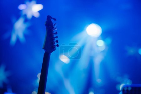 Kopfplatte einer Gitarre gegen eine blau beleuchtete Bühne mit sternförmigem Bokeh und Lichtstrahlen.