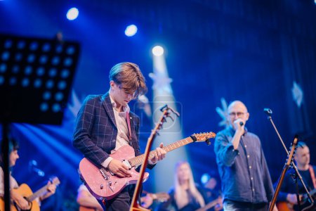 Photo for Valmiera, Latvia - December 28, 2023 -  young male guitarist in focus playing on stage with other musicians and a singer in the background, under blue stage lighting. - Royalty Free Image