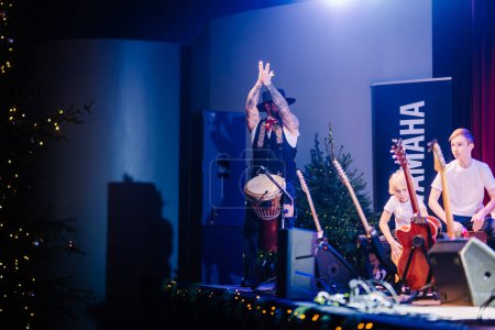 Téléchargez les photos : Valmiera, Lettonie - 28 décembre 2023 - musicien tatoué levant la main, jouant de la batterie sur scène avec des décorations de Noël, des guitares et un enfant à proximité. - en image libre de droit