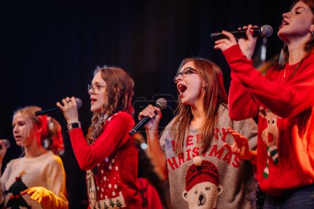 Téléchargez les photos : Valmiera, Lettonie - 28 décembre 2023 - groupe de personnes se produisant sur scène, portant des chandails de Noël festifs et chantant dans des microphones avec des instruments de musique en arrière-plan - en image libre de droit