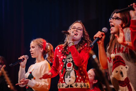 Téléchargez les photos : Valmiera, Lettonie - 28 décembre 2023 - groupe de personnes se produisant sur scène, portant des chandails de Noël festifs et chantant dans des microphones avec des instruments de musique en arrière-plan - en image libre de droit