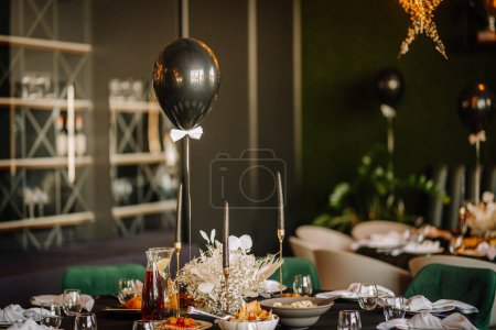 Foto de Riga, Letonia - 6 de diciembre de 2023 - La foto se centra en un globo negro con corbata de lazo, con un ambiente festivo de mesa con velas, flores blancas y vajilla elegante en el fondo. - Imagen libre de derechos