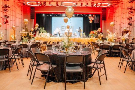 Foto de Sala de eventos festivos con mesas preparadas para una gala, manteles brillantes, globos dorados y un escenario con intérpretes en el fondo. - Imagen libre de derechos