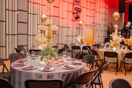 Foto de Lujosa configuración de eventos con candelabros y globos dorados, colocados contra un escenario con iluminación roja y paneles de pared estampados. - Imagen libre de derechos