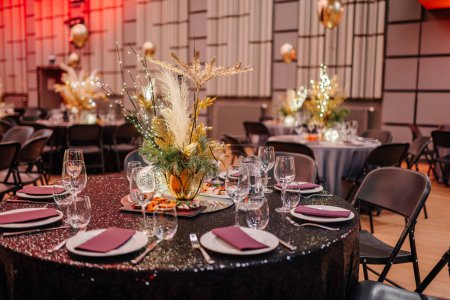 Foto de Mesa de eventos elegantemente establecida con un mantel brillante, centro de mesa floral de oro y vajilla arreglada, con mesas más decoradas en el fondo. - Imagen libre de derechos