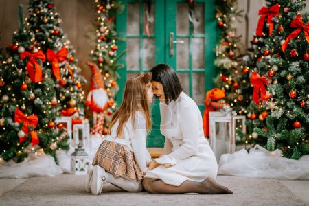 Photo for A tender moment between a mother and daughter, touching foreheads and smiling, with a Christmas tree backdrop. - Royalty Free Image