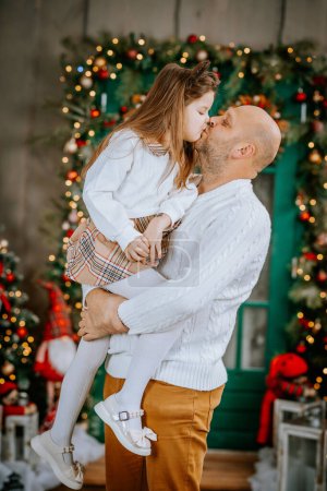 Photo for Father carrying his daughter, who is giving him a kiss, in a festive Christmas setting. - Royalty Free Image