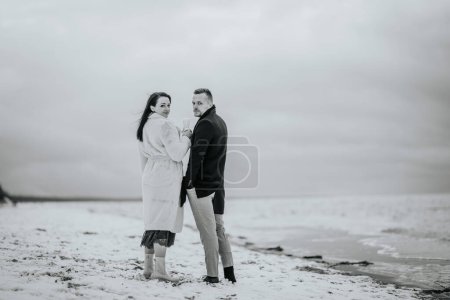 Photo for Monochrome shot of a couple standing on a snowy beach, the woman holding a champagne glass, both looking back at the camera. - Royalty Free Image