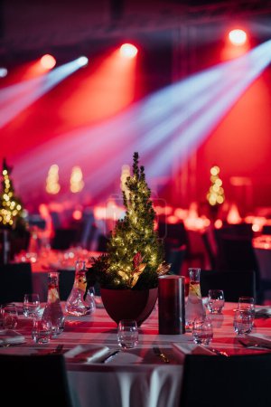 Foto de Centro del árbol de Navidad festivamente iluminado en una mesa de comedor con vajilla elegante, bajo iluminación de escenario roja y blanca. - Imagen libre de derechos