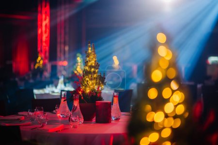 Foto de Mesa con una pieza central de árbol de Navidad iluminada en una sala de banquetes, con un efecto bokeh de las luces y la iluminación de escenario azul. - Imagen libre de derechos