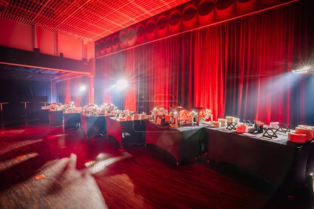 buffet setup in an event hall with serving dishes, under dramatic red drapery and intense stage lighting.