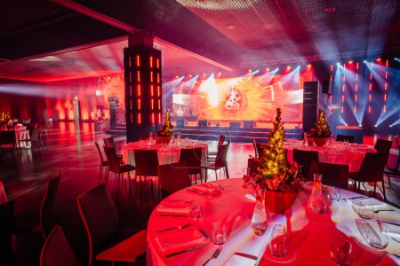 banquet hall with set tables, Christmas tree centerpieces, and a vibrant stage with red and blue lighting effects.