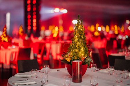 Foto de Mesa con una pieza central de árbol de Navidad iluminada en una sala de banquetes con iluminación ambiental roja y mesas de fondo borrosas. - Imagen libre de derechos