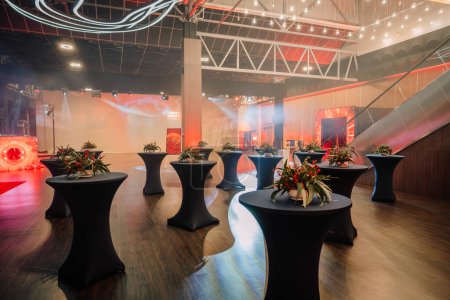Eleganter Veranstaltungsraum mit Cocktailtischen, Blumenarrangements, Ambientebeleuchtung und einer Bühne mit roter Hintergrundbeleuchtung.