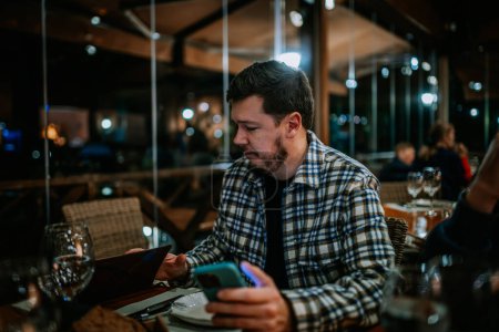 Foto de Málaga, España - 21 de enero de 2024 - hombre con camisa a cuadros mirando su smartphone en un restaurante con otros comensales al fondo. - Imagen libre de derechos