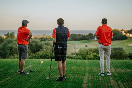 Téléchargez les photos : Malaga, Espagne - 21 janvier 2024 - Trois golfeurs sur un tee-box regardant vers un fairway menant à la mer, avec des arbres et un ciel dégagé. - en image libre de droit
