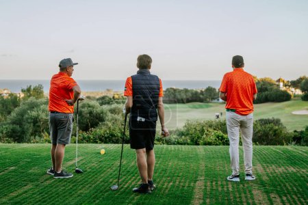Téléchargez les photos : Malaga, Espagne - 21 janvier 2024 - Trois golfeurs sur un tee-box regardant vers un fairway menant à la mer, avec des arbres et un ciel dégagé. - en image libre de droit