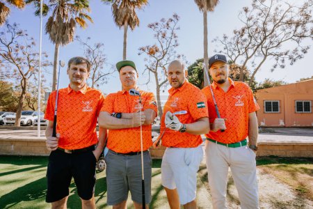 Téléchargez les photos : Malaga, Espagne - Janvier, 21, 2024 - Quatre golfeurs en chemises orange assorties, posant avec des clubs, devant un parking avec des palmiers. - en image libre de droit