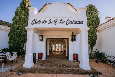 Foto de Málaga, España - 21 de enero de 2024 - Una escena de carretera soleada con una señal para "Club de Golf La Canada" a la izquierda, en medio de árboles y un cielo azul claro. - Imagen libre de derechos