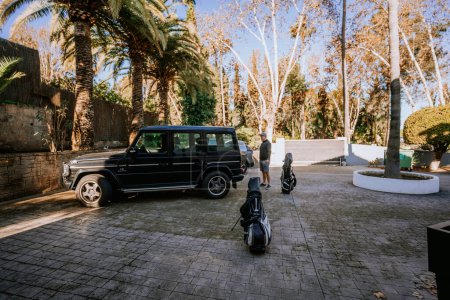 Téléchargez les photos : Malaga, Espagne - 21 janvier 2024 - personne debout à côté de sacs de golf garés sur une allée pavée avec un VUS noir et des palmiers en arrière-plan. - en image libre de droit