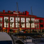 Sotogrande, Spain - January, 21, 2024 - The evening sun illuminates a red building with reflective windows at a marina lined with palm trees and moored boats in the foreground.