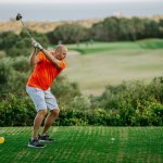 Malaga, Spain - January, 21, 2024 - A bald man in an orange shirt and white shorts is in the middle of a golf swing on a course with a sea view.