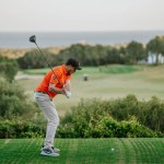 Malaga, Spain - January, 21, 2024 -  A golfer in an orange shirt is mid-swing on a golf course with a backdrop of the sea and a clear sky.