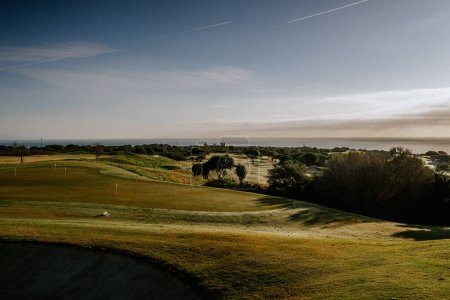 Foto de Sotogrande, España - 23 de enero de 2024 - Una vista panorámica de un campo de golf con calles onduladas, búnkeres y vistas distantes del mar y el horizonte bajo un cielo con pistas de avión. - Imagen libre de derechos